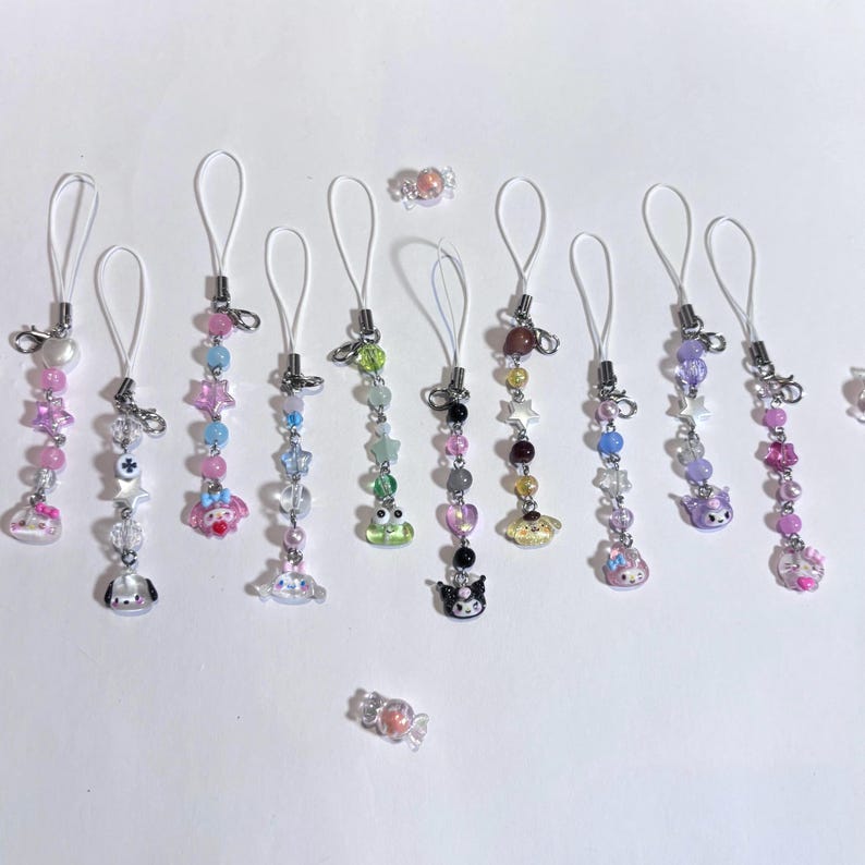 May include: A collection of beaded phone charms in various colors, featuring charms with cartoon characters, stars, and hearts. Each charm has a white loop and silver-colored metal accents. Two small candy-shaped charms are also present.