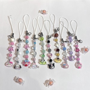May include: A collection of colorful beaded phone charms with various charms, including stars, hearts, and cartoon characters. The charms feature pink, blue, green, and purple beads, with silver-toned metal accents and white cords. Several candies are also scattered around.