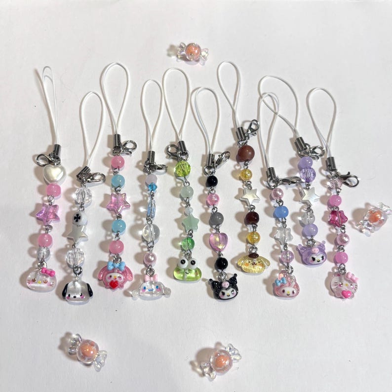 May include: A collection of colorful beaded phone charms with various charms, including stars, hearts, and cartoon characters. The charms feature pink, blue, green, and purple beads, with silver-toned metal accents and white cords. Several candies are also scattered around.