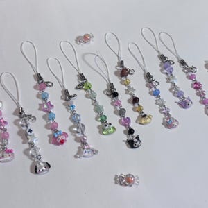 May include: A collection of beaded phone charms in various colors, featuring star, heart, and character charms. Each charm has a white loop for attachment. Scattered candies are also visible.