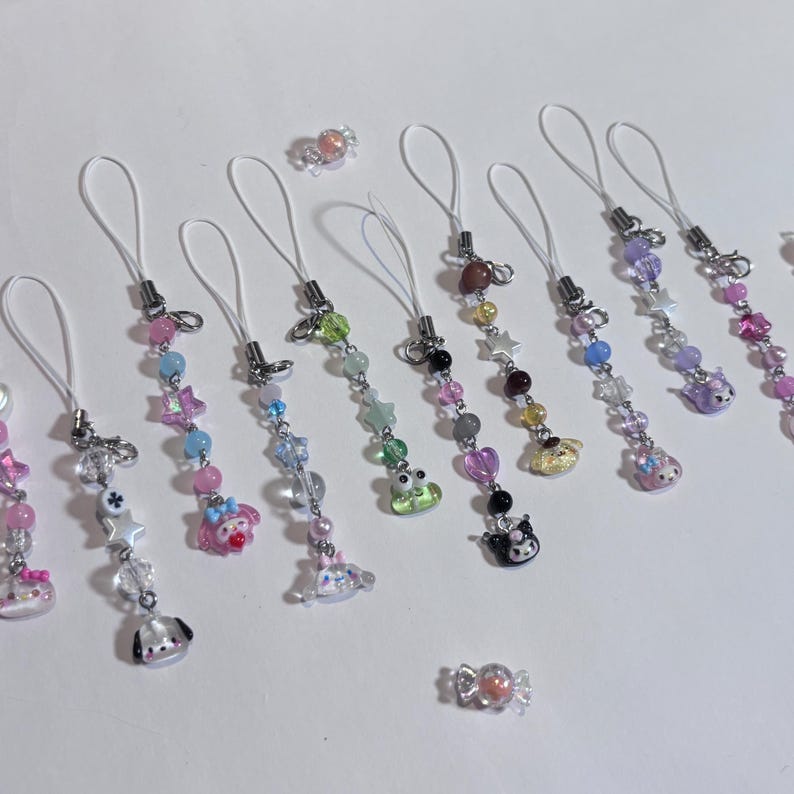 May include: A collection of beaded phone charms in various colors, featuring star, heart, and character charms. Each charm has a white loop for attachment. Scattered candies are also visible.