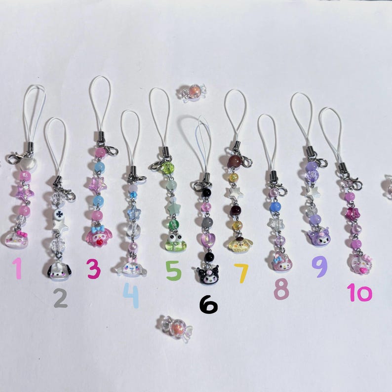 May include: A collection of ten beaded phone charms in various colors, each with a different charm at the bottom. Charms include stars, hearts, and cartoon characters. Each charm has a silver-colored clasp and loop.