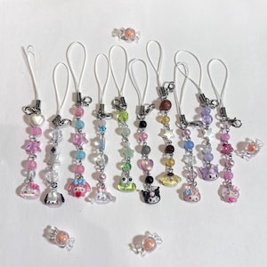 May include: A collection of beaded phone charms with various designs, including stars, hearts, and cartoon characters. The charms feature a mix of colorful beads and clear accents, with a white loop for attachment. Several candy-shaped charms are also scattered around.
