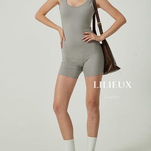 Puede incluir: Un mono gris sin mangas con cuello redondo. La modelo lleva calcetines blancos y zapatillas blancas. La modelo lleva un bolso de cuero marrón.