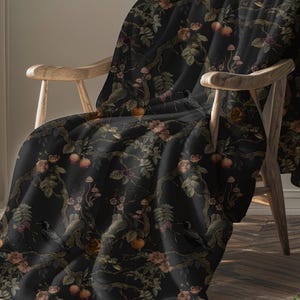 Puede incluir: Una manta negra con un diseño botánico que presenta rosas, setas y frutas en tonos melocotón, verde y marrón. El diseño incluye imágenes de cuervos, sobre una silla de madera.