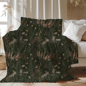 May include: A dark green throw blanket featuring a botanical design of flowers and foliage in shades of brown, beige, and yellow. The blanket is draped over a white sofa. The blanket's design features a detailed floral print, creating a cosy atmosphere.