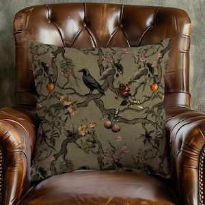 May include: Square decorative pillow with a botanical design featuring a black crow perched on a tree branch. The pillow has a muted green background with illustrations of flowers, fruit, and mushrooms. The pillow is placed on a brown leather armchair.