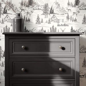 Puede incluir: Una c&oacute;moda gris oscuro con tres cajones y pomos plateados. La c&oacute;moda est&aacute; colocada contra un papel pintado que representa una escena de monta&ntilde;a y bosque en blanco y negro con caba&ntilde;as y ciervos. Una jarr&oacute;n gris con ramas est&aacute; encima.