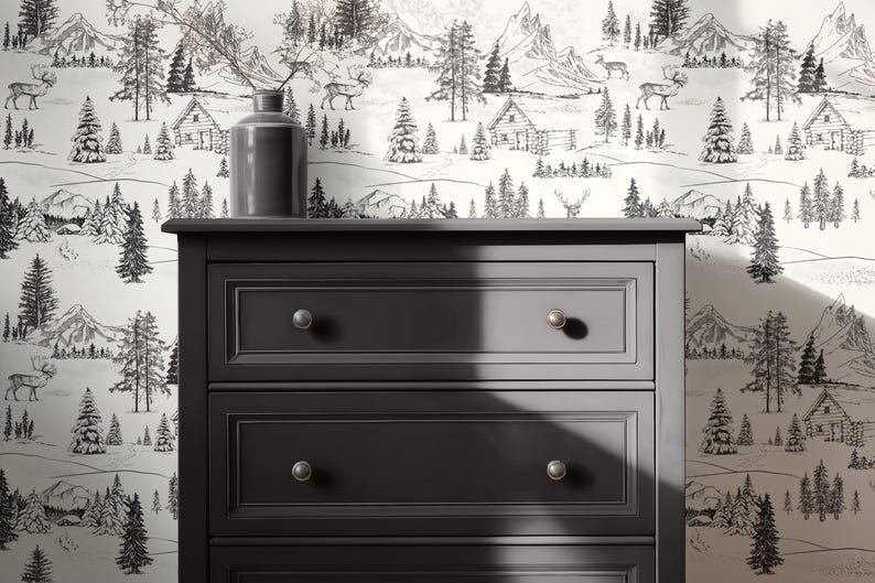 Puede incluir: Una c&oacute;moda gris oscuro con tres cajones y pomos plateados. La c&oacute;moda est&aacute; colocada contra un papel pintado que representa una escena de monta&ntilde;a y bosque en blanco y negro con caba&ntilde;as y ciervos. Una jarr&oacute;n gris con ramas est&aacute; encima.