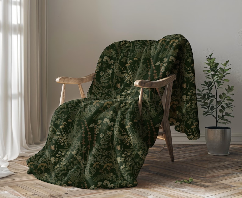 Puede incluir: Una manta verde oscuro con un estampado floral sobre una silla de madera. La manta presenta un dise&ntilde;o repetido de flores y follaje verde claro y beige. Una peque&ntilde;a planta en maceta est&aacute; a la derecha de la silla.