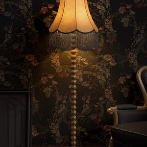 May include: A tall, wooden floor lamp with a beige lampshade and dark blue fringe. The lamp has a beaded stem and sits on a round base. The background features floral wallpaper and a dark-colored chair.