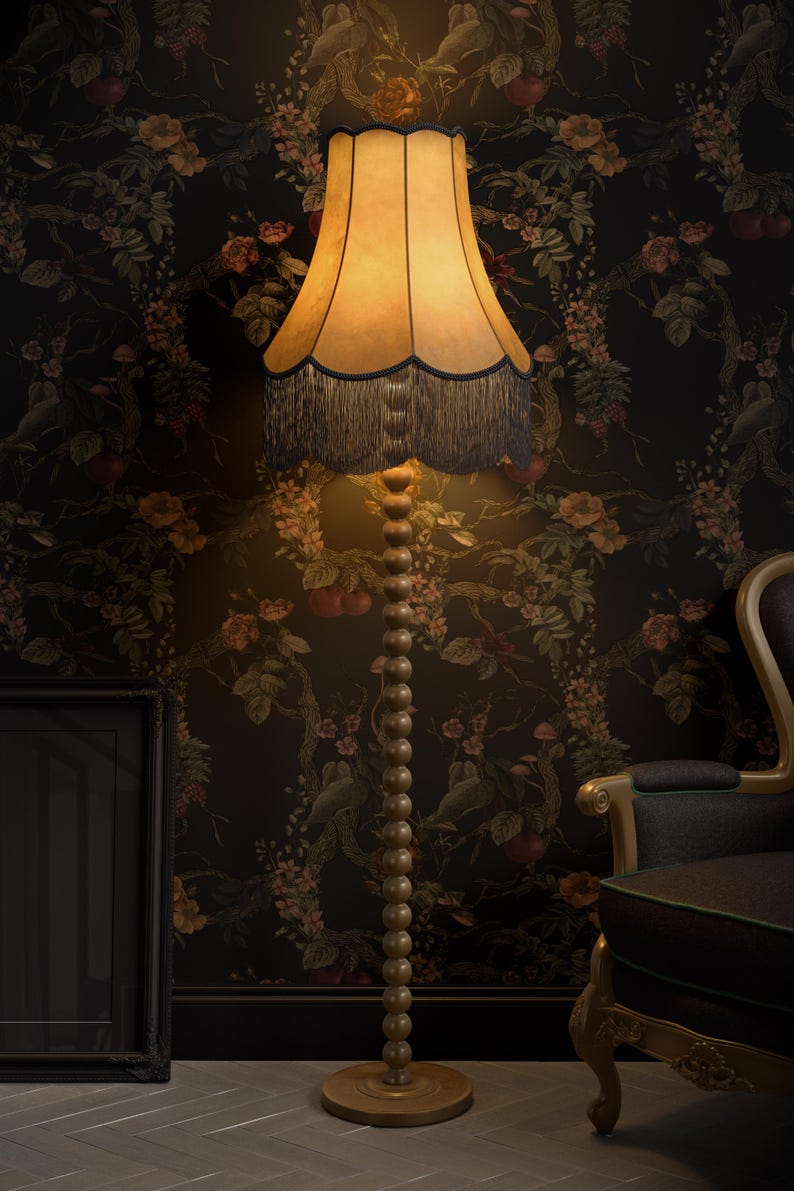 May include: A tall, wooden floor lamp with a beige lampshade and dark blue fringe. The lamp has a beaded stem and sits on a round base. The background features floral wallpaper and a dark-colored chair.