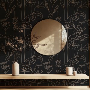 Puede incluir: Un papel tapiz negro y dorado con un dise&ntilde;o floral minimalista. El papel tapiz presenta delicados dibujos lineales de flores y hojas. Un espejo redondo y dos jarrones de cer&aacute;mica se colocan en un estante de madera frente al papel tapiz.