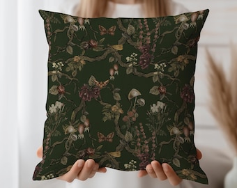 May include: A square decorative pillow with a dark green background and a botanical pattern. The pattern includes illustrations of mushrooms, flowers, butterflies, and branches in shades of brown, burgundy, and cream.