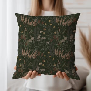 Könnte beinhalten: Ein quadratisches Zierkissen mit dunkelgrünem Hintergrund und einem sich wiederholenden Blumenmuster. Das Muster umfasst verschiedene Pflanzen in Braun-, Beige- und Gelbtönen. Das Kissen wird von einer Person gehalten.