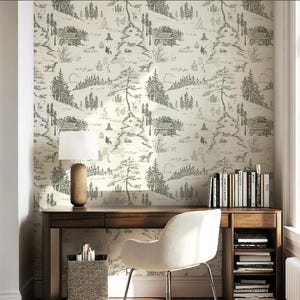 May include: A home office with a wooden desk, a white chair, and a lamp. The wall is covered in a cream-colored wallpaper with a black and white forest scene. Books are on the desk and in a shelf below.