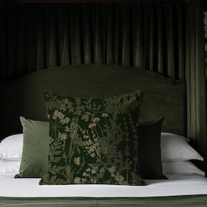 May include: A dark green bed with a velvet headboard and matching curtains. The bed is made with white sheets and several pillows. The pillows include a floral patterned pillow and two solid green pillows. A lamp and framed botanical prints are visible.