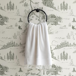 May include: A white, folded towel hangs from a black metal ring. The towel has a fringed edge and two embroidered lines near the bottom. The background is a wallpaper with a forest scene in shades of gray.