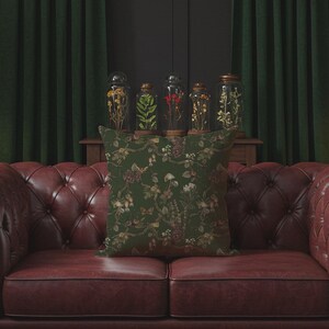 May include: A decorative square pillow with a dark green background and a floral pattern in shades of brown and cream. The pillow is placed on a red leather sofa. Behind the sofa are glass jars with dried plants and framed botanical prints.
