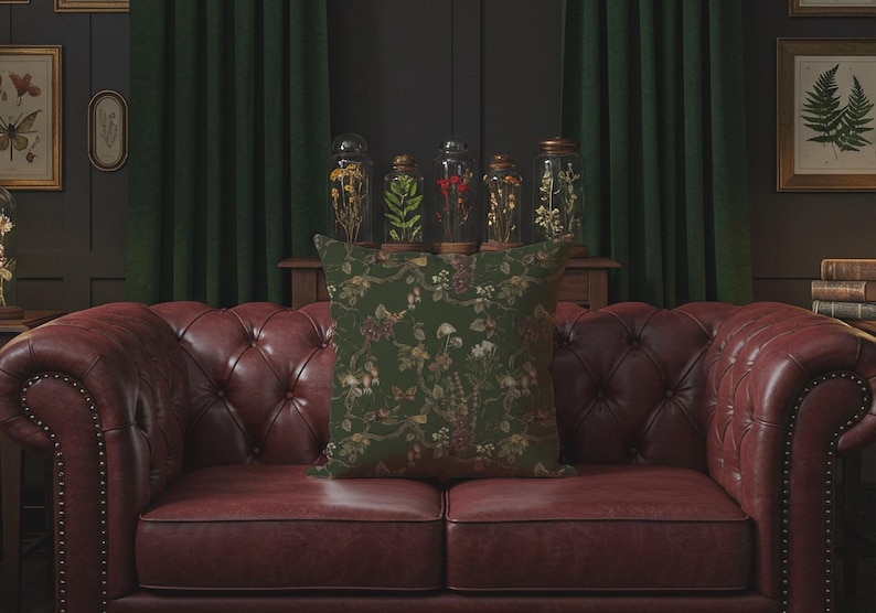 May include: A decorative square pillow with a dark green background and a floral pattern in shades of brown and cream. The pillow is placed on a red leather sofa. Behind the sofa are glass jars with dried plants and framed botanical prints.