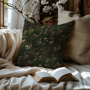 May include: A decorative throw pillow with a dark green background and a floral pattern featuring mushrooms, berries, and flowers in shades of burgundy, cream, and brown. The pillow is placed on a cream-colored blanket.