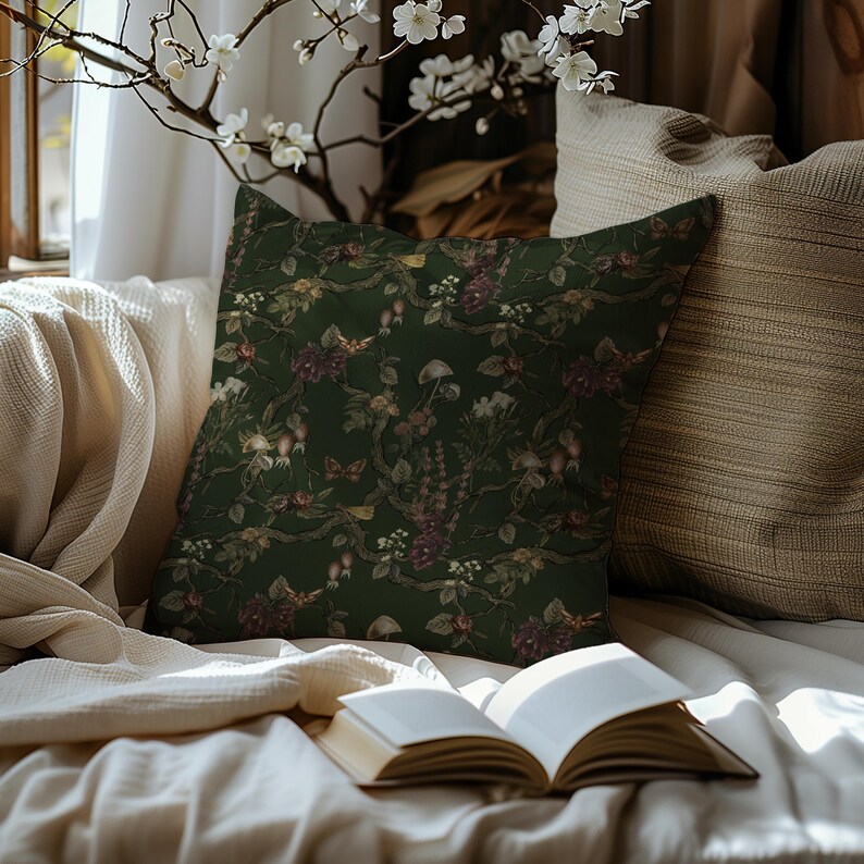 May include: A decorative throw pillow with a dark green background and a floral pattern featuring mushrooms, berries, and flowers in shades of burgundy, cream, and brown. The pillow is placed on a cream-colored blanket.
