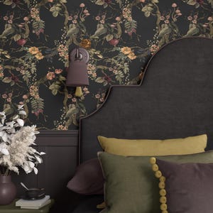 May include: A bedroom scene featuring a dark floral wallpaper with botanical illustrations. The bed has a dark headboard and is adorned with pillows in shades of green, yellow, and purple. A wall sconce and a small side table with a vase and books complete the decor.
