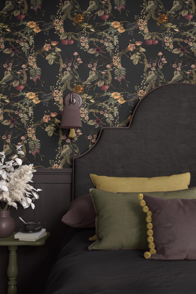 May include: A bedroom scene featuring a dark floral wallpaper with botanical illustrations. The bed has a dark headboard and is adorned with pillows in shades of green, yellow, and purple. A wall sconce and a small side table with a vase and books complete the decor.