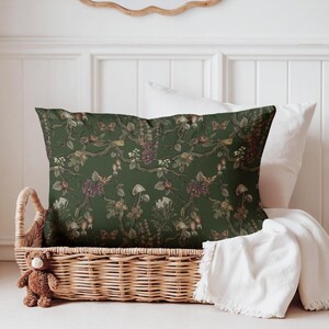 May include: A rectangular decorative pillow with a dark green background and a botanical pattern. The pattern includes flowers, mushrooms, and butterflies in shades of burgundy, brown, and cream. The pillow is placed in a wicker basket with a teddy bear.