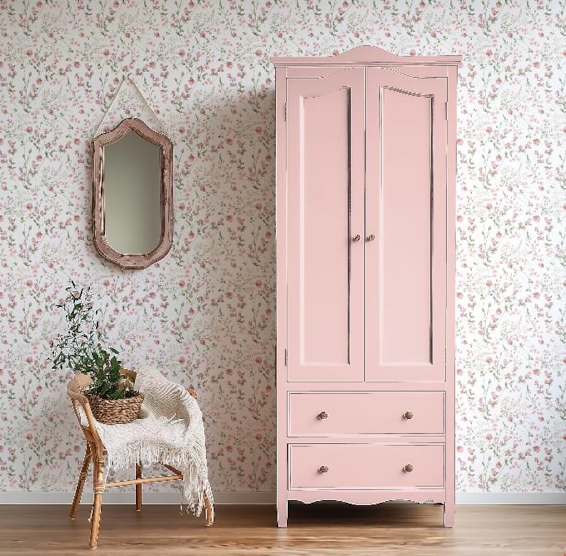 Op de afbeelding: Een lichtroze houten kledingkast met twee deuren en twee lades. De kledingkast heeft een klassiek ontwerp met afgeronde randen en messing knoppen. De achtergrond toont bloemenbehang, een decoratieve spiegel en een rotan stoel.