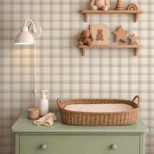 Puede incluir: Una habitación infantil con una cómoda verde, una cesta para cambiar pañales de mimbre y estantes de madera. Los estantes exhiben un oso de peluche, un conejito, un libro y estrellas de madera. También se ve una lámpara blanca y un papel pintado a cuadros.