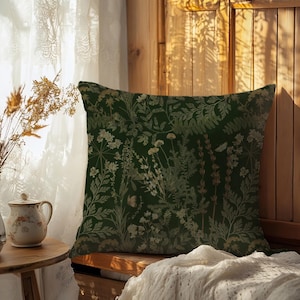 May include: A decorative square pillow with a dark green background and a botanical pattern in shades of beige and light green. The pillow is placed on a wooden bench near a window with sheer curtains. A small table with a vase and a cup is in the foreground.