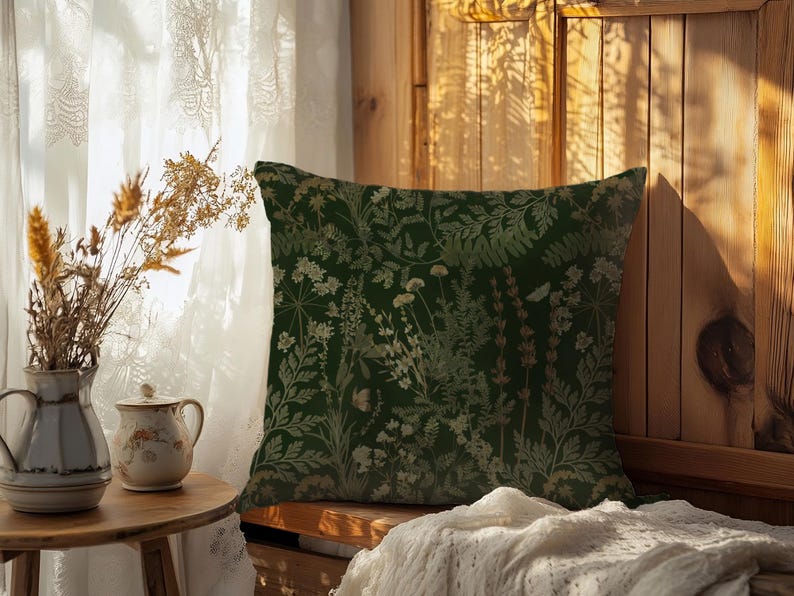May include: A decorative square pillow with a dark green background and a botanical pattern in shades of beige and light green. The pillow is placed on a wooden bench near a window with sheer curtains. A small table with a vase and a cup is in the foreground.