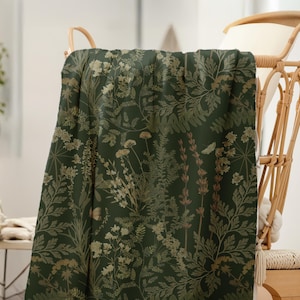 Puede incluir: Una manta verde oscuro con un estampado bot&aacute;nico de varias flores y follaje en tonos dorados y marrones. La manta est&aacute; sobre una silla de mimbre, mostrando el detallado dise&ntilde;o floral.