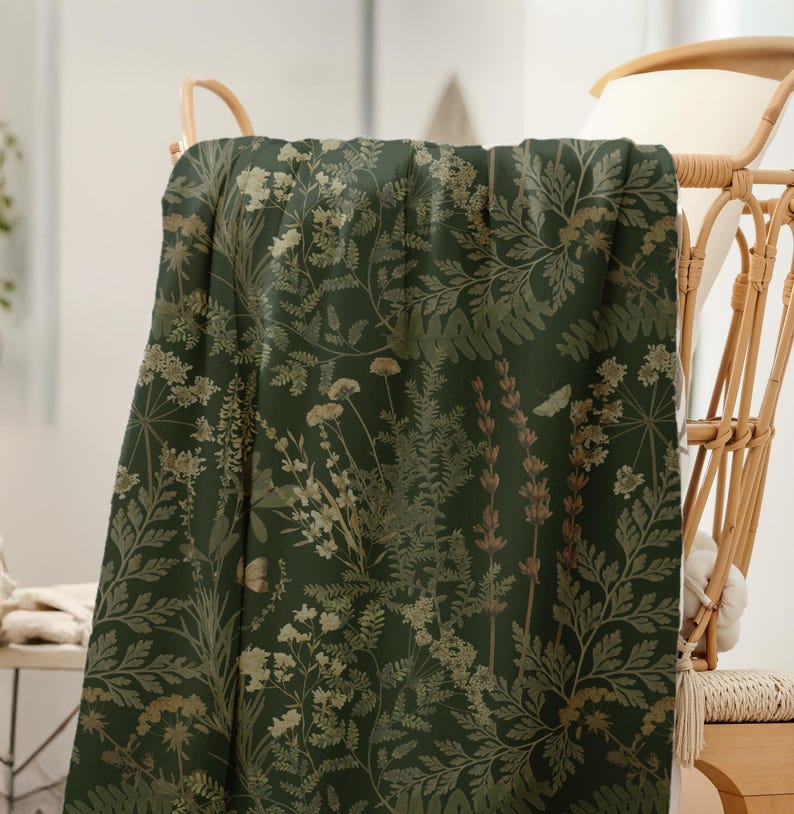 Puede incluir: Una manta verde oscuro con un estampado bot&aacute;nico de varias flores y follaje en tonos dorados y marrones. La manta est&aacute; sobre una silla de mimbre, mostrando el detallado dise&ntilde;o floral.