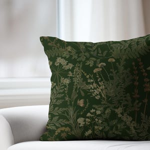 May include: A square decorative pillow with a dark green background and a botanical pattern. The pattern features various plants in shades of gold and beige, including ferns, flowers, and other foliage. The pillow is placed on a white sofa.