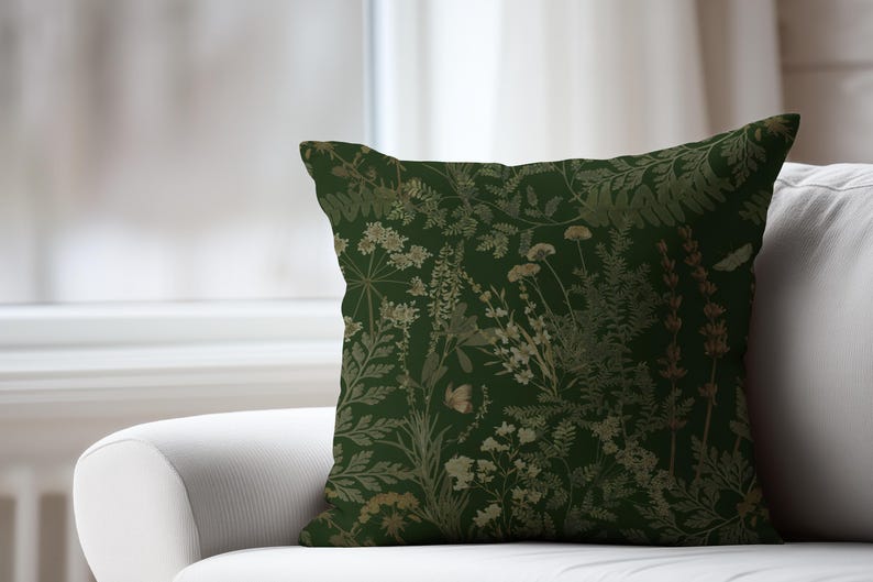 May include: A square decorative pillow with a dark green background and a botanical pattern. The pattern features various plants in shades of gold and beige, including ferns, flowers, and other foliage. The pillow is placed on a white sofa.