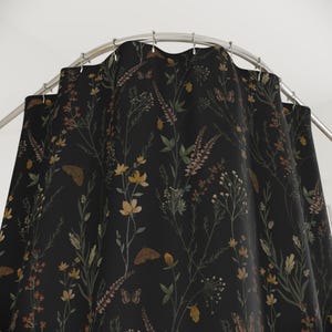 May include: A black shower curtain with a botanical design. The pattern features various wildflowers, butterflies, and foliage in shades of yellow, brown, and green. The curtain hangs from a curved, silver-colored shower rod.