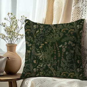 May include: A square decorative pillow with a dark green background and a botanical pattern of various plants in shades of green, beige, and brown. The pillow is placed on a white surface near a vase with dried flowers.