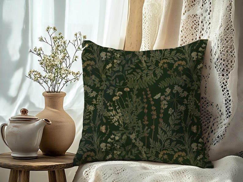 May include: A square decorative pillow with a dark green background and a botanical pattern of various plants in shades of green, beige, and brown. The pillow is placed on a white surface near a vase with dried flowers.