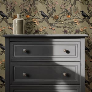 May include: A dark gray dresser with three drawers and silver knobs. A light-colored ceramic vase sits on top of the dresser. The background is a wallpaper with a botanical design featuring birds, flowers, and fruit.