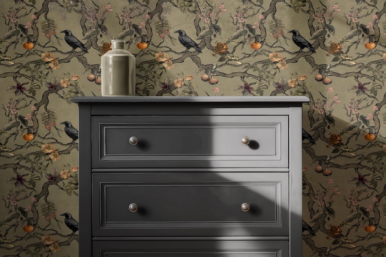 May include: A dark gray dresser with three drawers and silver knobs. A light-colored ceramic vase sits on top of the dresser. The background is a wallpaper with a botanical design featuring birds, flowers, and fruit.