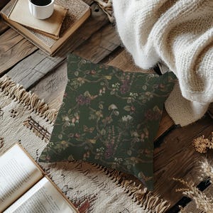 May include: A square decorative pillow with a dark green background and a floral pattern. The pattern includes branches, flowers, and mushrooms in shades of burgundy, cream, and brown. The pillow is placed on a wooden surface.