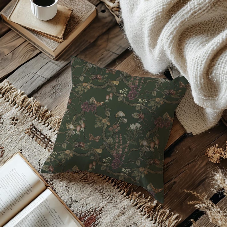 May include: A square decorative pillow with a dark green background and a floral pattern. The pattern includes branches, flowers, and mushrooms in shades of burgundy, cream, and brown. The pillow is placed on a wooden surface.