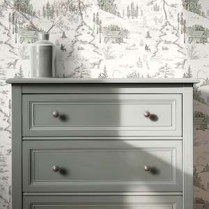 May include: A gray dresser with three drawers and silver knobs. A gray vase with branches sits on top of the dresser. The background features a black and white forest-themed wallpaper.