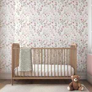 Op de afbeelding: Een houten wieg met een witte matras en een grijze deken in een kinderkamer. De muren zijn bedekt met bloemenbehang met roze en groene accenten. Een bruine teddybeer zit op de vloer voor de wieg.