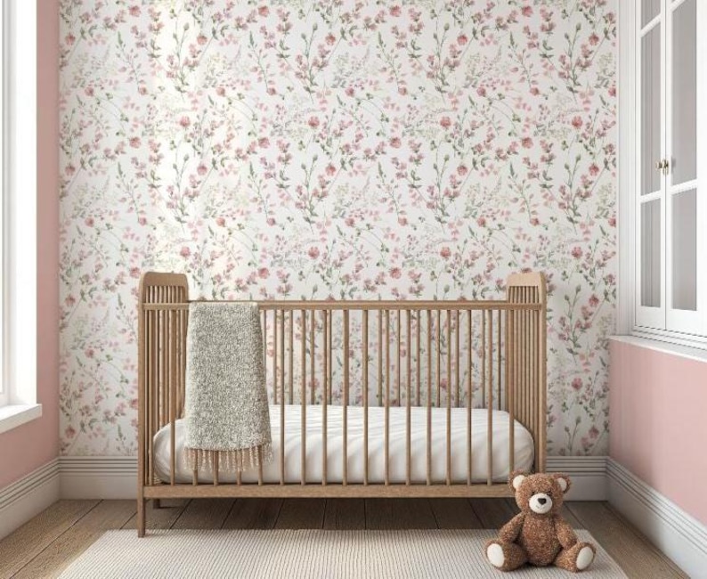 Op de afbeelding: Een houten wieg met een witte matras en een grijze deken in een kinderkamer. De muren zijn bedekt met bloemenbehang met roze en groene accenten. Een bruine teddybeer zit op de vloer voor de wieg.
