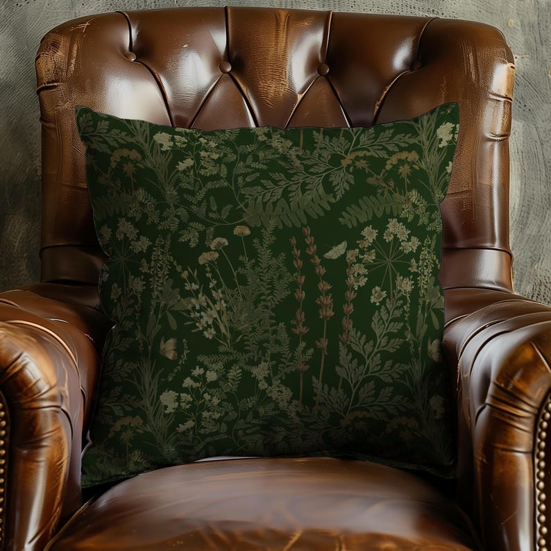 May include: Square decorative pillow with a dark green background and a botanical pattern in shades of beige and brown. The pillow is placed on a brown leather armchair.