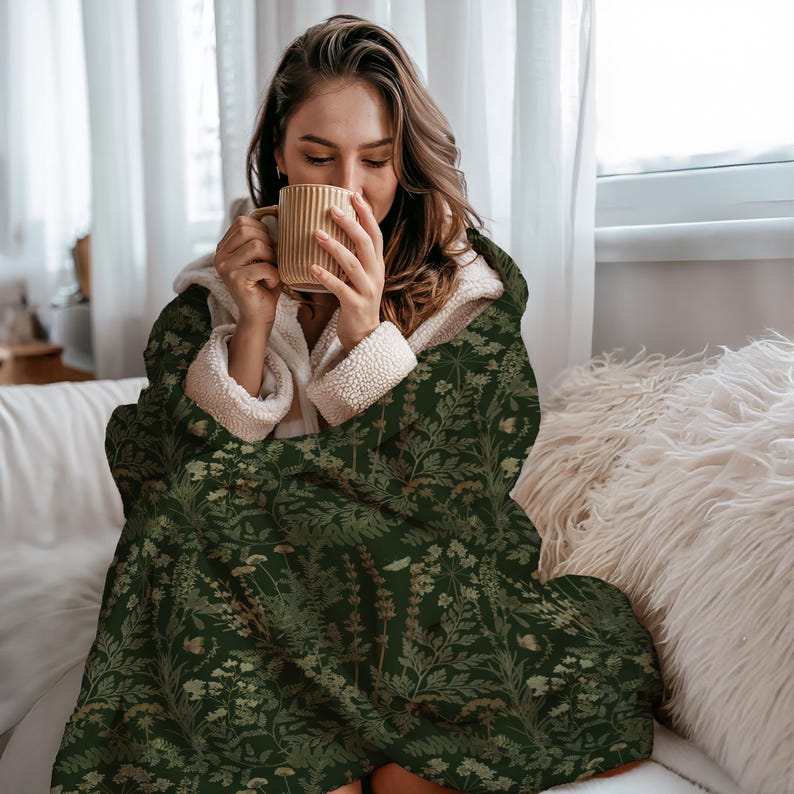 Puede incluir: Una manta con capucha con un estampado bot&aacute;nico en tonos verdes y beige. La manta tiene un forro de sherpa color crema y est&aacute; envuelta alrededor de una persona que sostiene una taza beige. La manta presenta un dise&ntilde;o floral detallado.