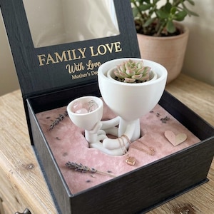 May include: A black box with a clear lid, featuring the text "FAMILY LOVE With Love Mother's Day" in gold lettering. Inside, a white ceramic figure holds a succulent and a small bowl with pink stones. A gold heart necklace and a pink heart-shaped decoration are also present.